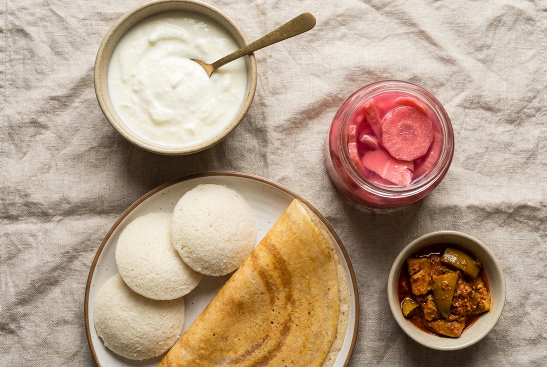 Overhead flat lay of Indian fermented foods — dahi, idli, dosa, kanji and pickle on a linen cloth