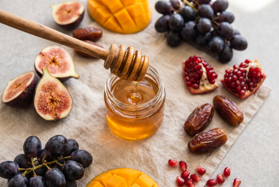 Overhead flat lay of honey being drizzled into a jar surrounded by fresh figs, mango, grapes, dates, and pomegranate