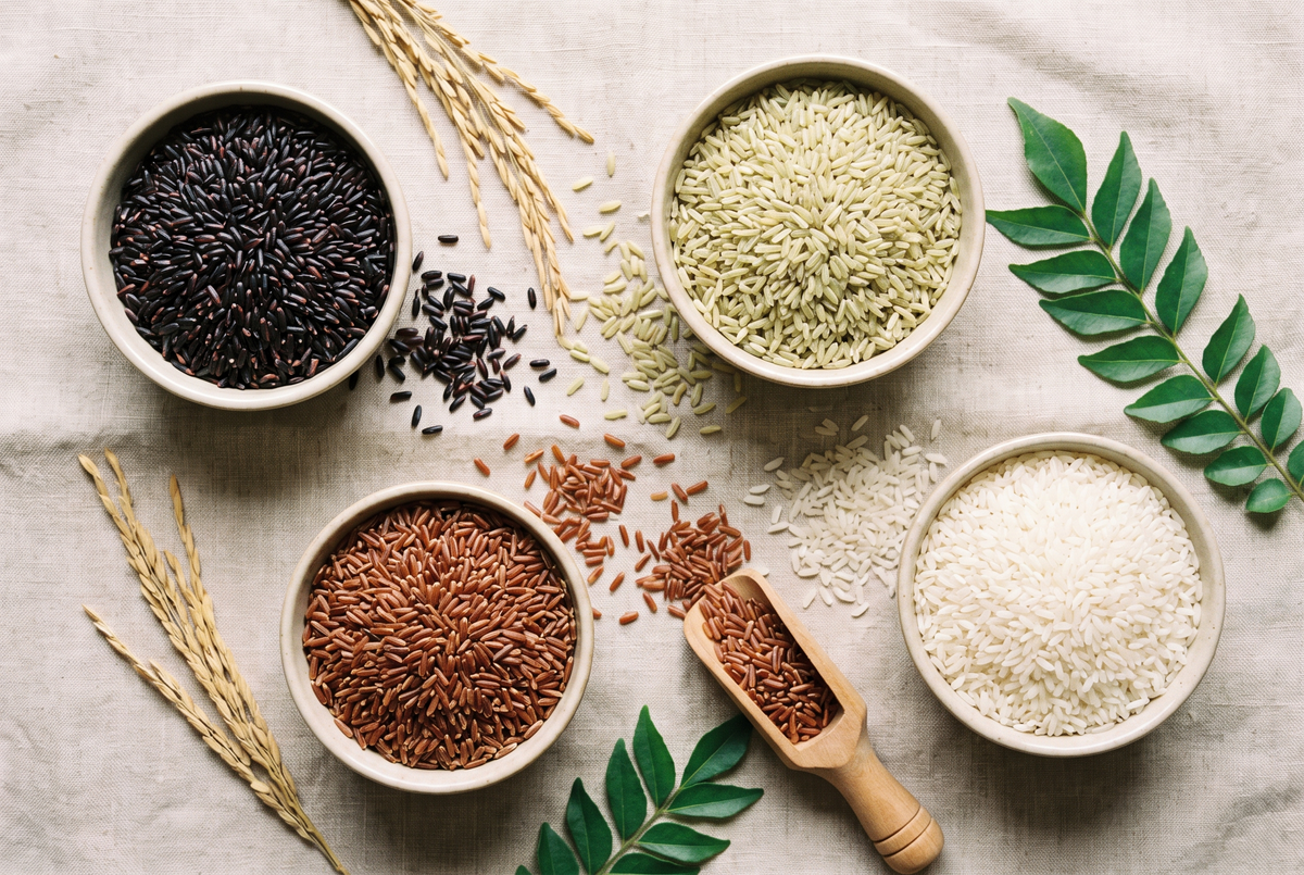 Overhead view of bowls of black, green, red and white rice on linen with rice stalks and curry leaves